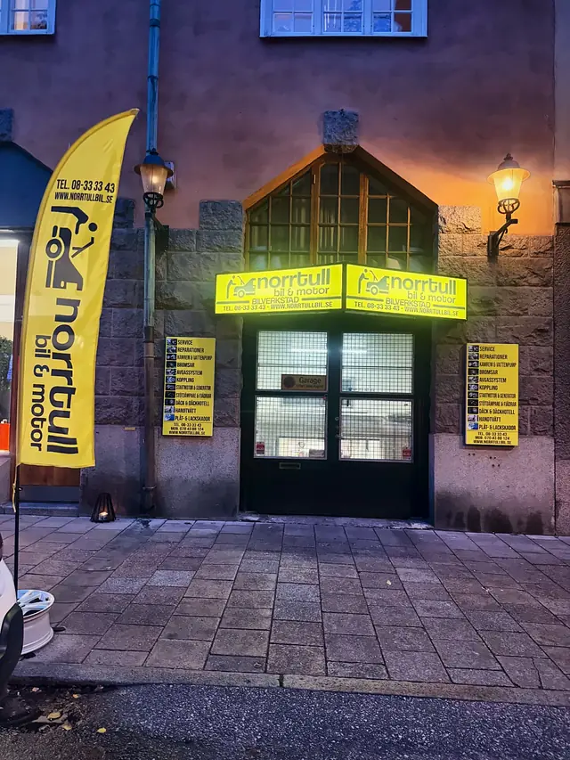 Entrén till Norrtull Bil & Motor på Norrtullsgatan 51 i kvällsljus med tänd gul skylt, gatlyktor och reklamflagga